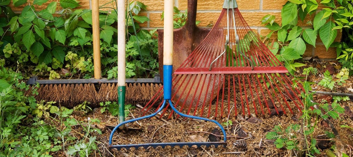 Close up of various types of rakes leaned against a brick wall outside.